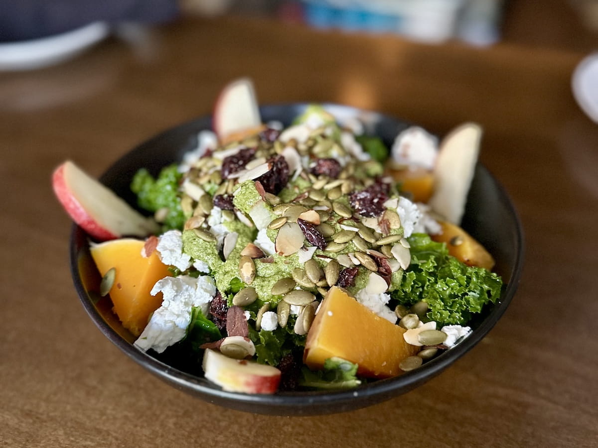 A close-up food photo of a vibrant autumn harvest salad served in a dark ceramic bowl at The Print House in Fleischmanns, NY, in the Catskills. The salad features dark leafy greens tossed with roasted squash chunks, sliced red apples, crumbled goat cheese, dried cranberries, slivered almonds, pepitas, and a green pesto dressing drizzled over the top.