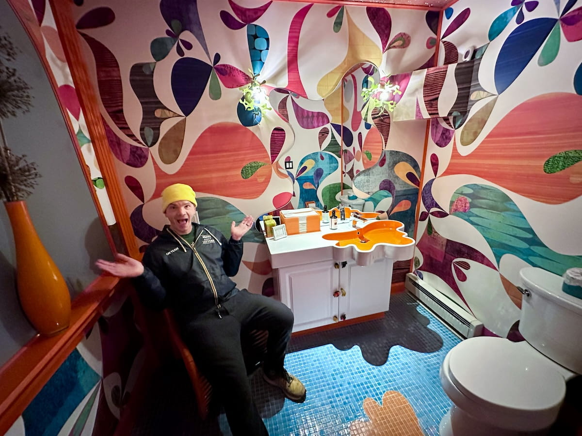 A wide interior shot of a whimsical, vibrantly decorated bathroom at The Roxbury Motel, featuring bold custom wallpaper with swirling retro patterns in pink, orange, teal, and purple. On the left, a man wearing a yellow beanie, dark jacket, and sweatpants sits with an excited expression and open hands. The bathroom includes a custom splash-shaped orange sink vanity, matching splash-shaped tile designs inlaid into the blue mosaic tile floor, an arched vanity mirror reflecting the colorful walls, and a white toilet on the right.