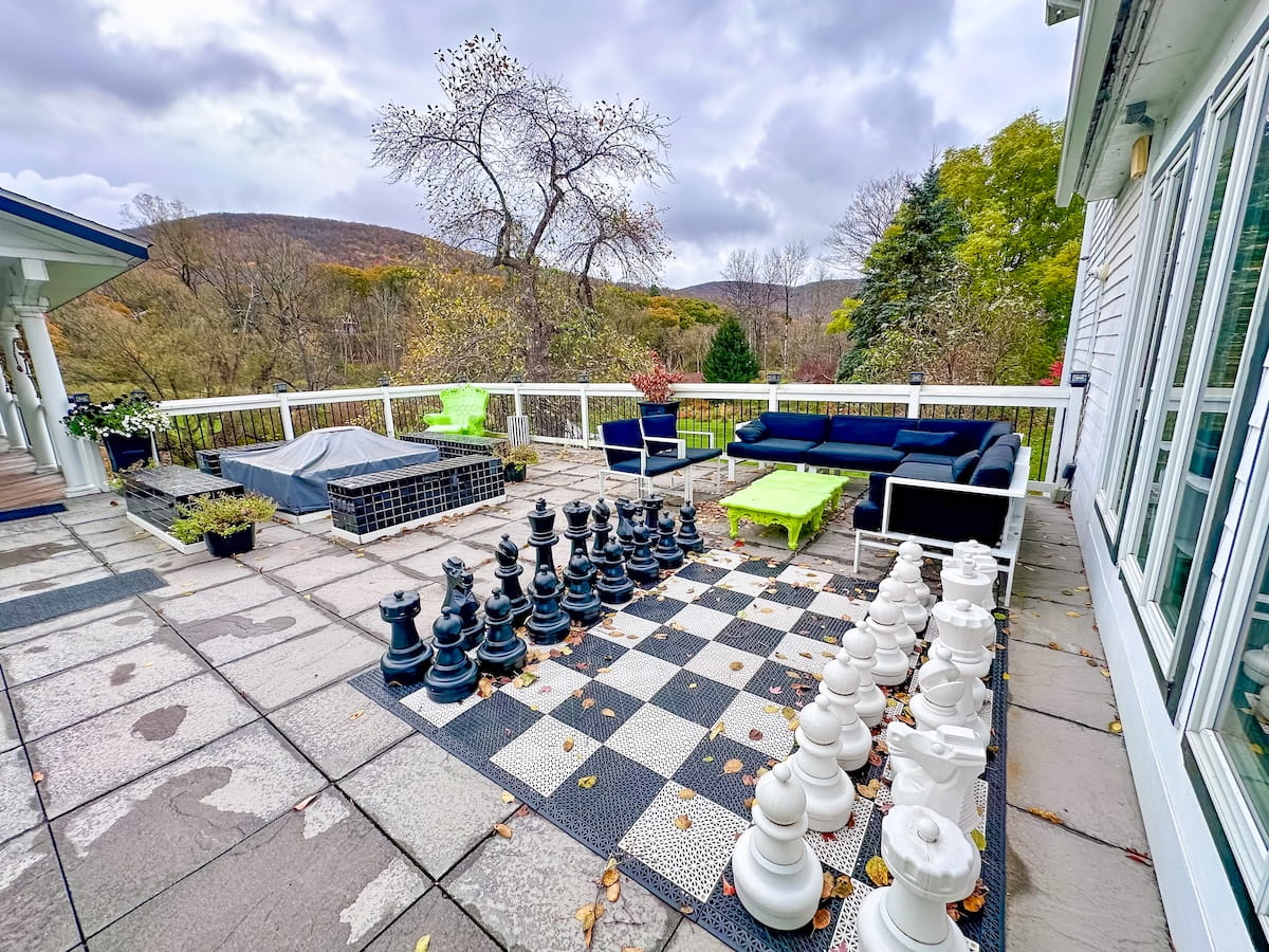 An outdoor stone patio at The Roxbury Motel featuring an oversized black-and-white chess set on a patterned ground board lined with large game pieces. In the background, modern outdoor lounge furniture includes a navy sectional sofa, neon-green accent tables, and a tiled fire pit set against a white railing overlooking autumn forest trees and distant Catskill mountains under an overcast sky.