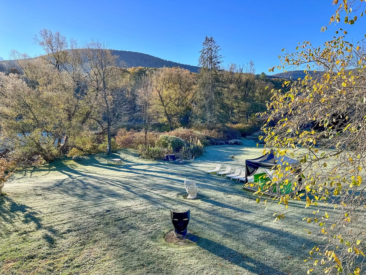 A high-angle landscape view looking out over the frost-covered grass lawn at The Roxbury Motel on a clear, sunny autumn morning. Two large face-shaped sculpture chairs—one white and one black—rest on the lawn near lounge chairs and a covered cabana. Surrounding the property are vibrant golden autumn trees, rolling mountain ridges, and a bright blue sky.