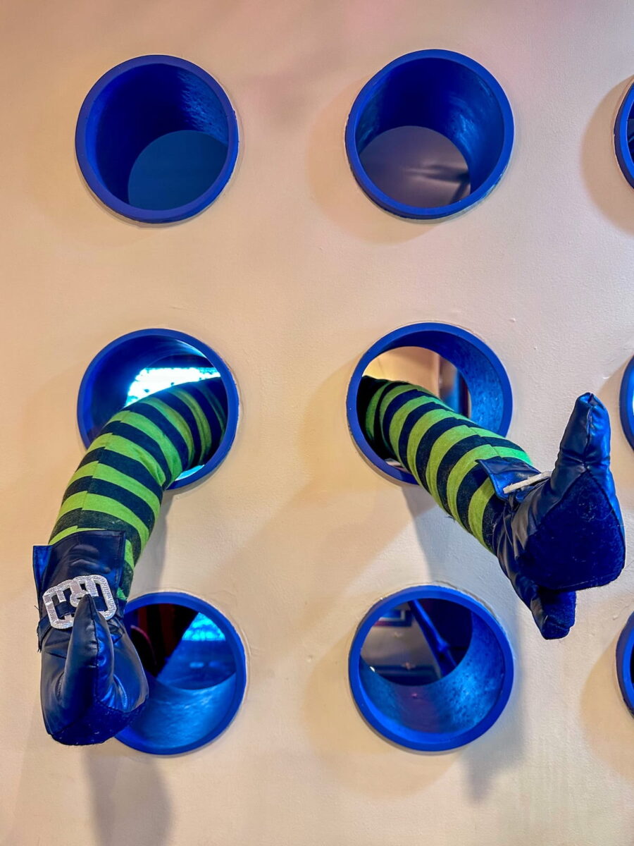 A close-up shot of a cream-colored wall featuring a grid of round blue porthole-style openings, with two green-and-black striped witch legs protruding outward from separate holes as part of Halloween decor at The Roxbury Motel. The legs wear black pointed witch shoes with silver buckle accents.