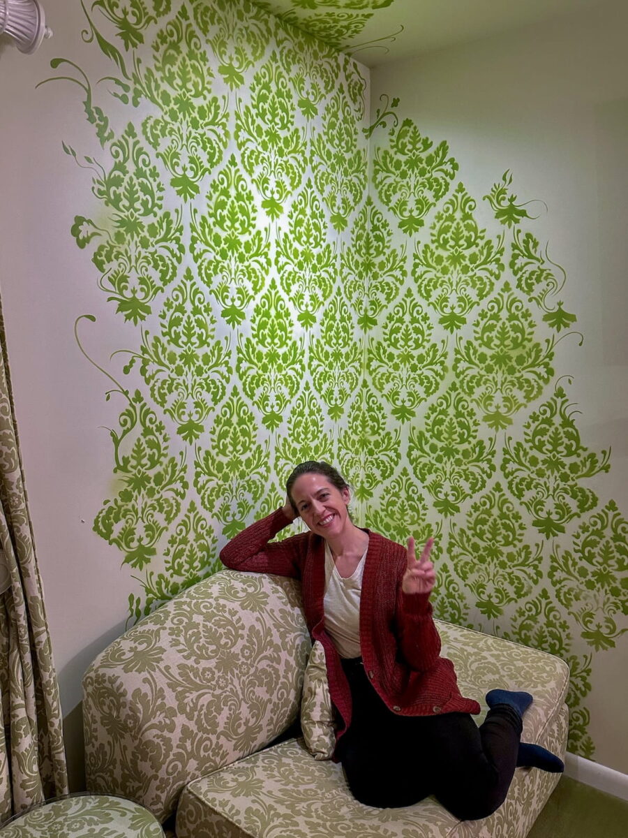 A vertical portrait of a smiling woman lounging on an upholstered chaise sofa inside the Maria's Curtains room at The Roxbury Motel. She wears a burgundy knit cardigan over a white shirt, black pants, and dark socks, resting her head against one hand while holding up a peace sign with the other. Behind her, the corner walls and ceiling feature intricate bright lime-green damask floral stencil patterns painted against a white wall, matching the patterned fabric of the sofa.