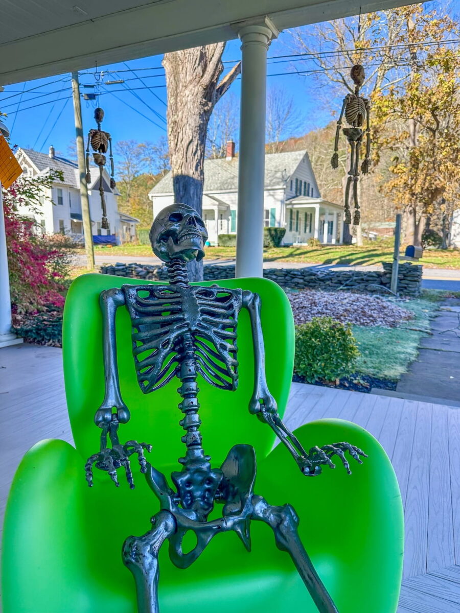 A Halloween display on the covered wooden porch of The Roxbury Motel featuring a metallic grey skeleton seated upright in a bright lime-green plastic armchair. In the background, additional smaller skeletons hang suspended from the porch ceiling, with white historic village houses and autumn trees visible across the street under a clear blue sky.