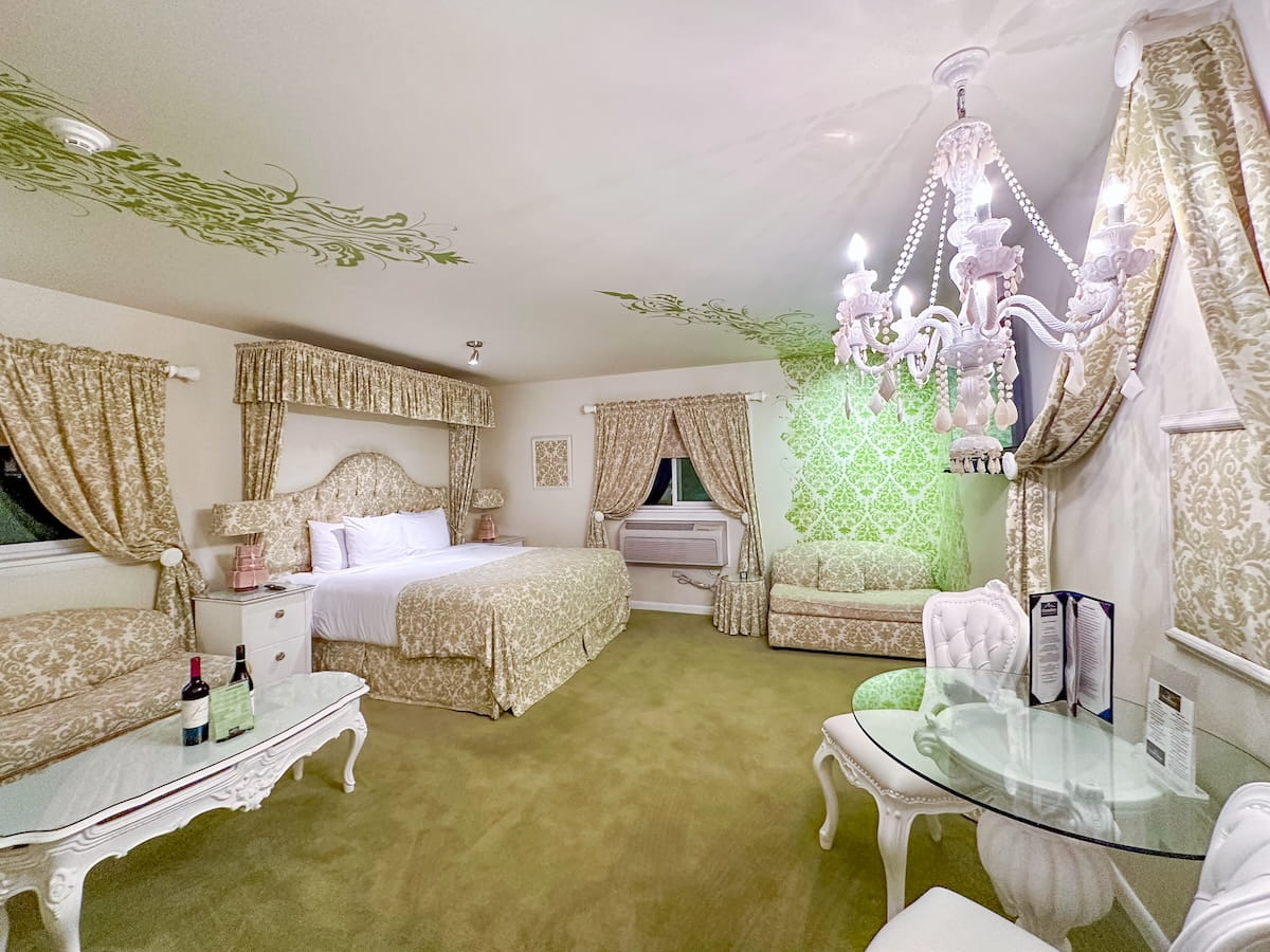 A wide view of the interior of the "Maria's Curtains" themed room at The Roxbury Motel, inspired by The Sound of Music. The room features plush green carpeting, green floral stenciling across the ceiling, matching gold-and-cream brocade fabrics on the bed canopy, drapes, and sofas, and an ornate white chandelier hanging above a glass-topped dining table with white tufted chairs.