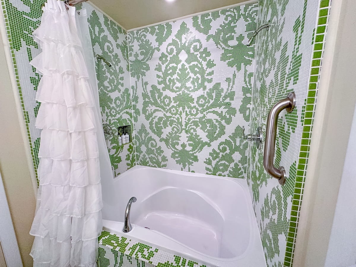 A close-up view of a custom-tiled bathroom inside the "Maria's Curtains" themed room at The Roxbury Motel. The deep white soaking tub and surrounding walls are lined with a striking green and white damask mosaic pattern, featuring a white ruffled shower curtain on the left, chrome fixtures, and a recessed shower niche with toiletry bottles on the back wall.