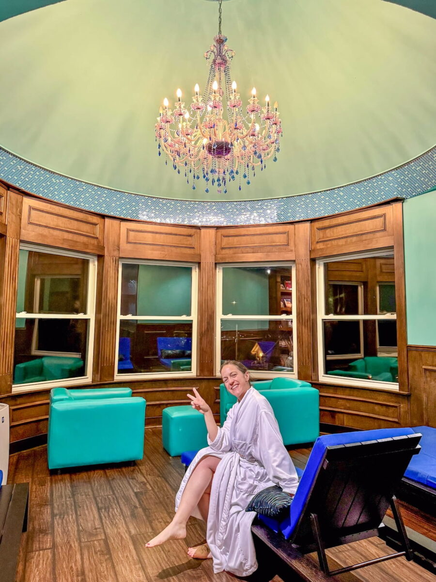 A smiling woman wearing a white plush robe sitting on a lounge chair and flashing a peace sign inside the Shimmer Spa at The Roxbury Motel. The circular room features rich wood paneling, large windows, bright turquoise armchairs, and a soft green domed ceiling centered by an ornate crystal chandelier with purple and blue accents.