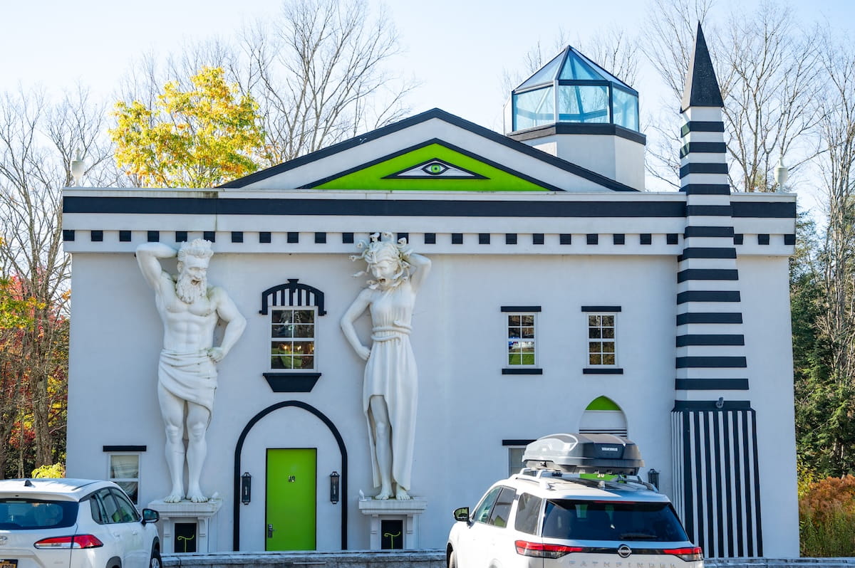 A front view of a whimsical, themed cottage building at The Roxbury at Stratton Falls in the Catskills. The white structure features bold black trim, a vibrant lime-green door, a roof gable accented with a stylized green-and-black eye motif, a glass cupola, a striped black-and-white obelisk tower, and two large white statues of mythological figures built into the facade flanking a window.