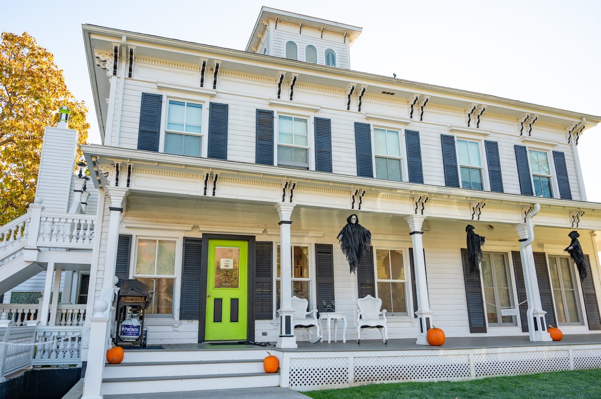 A front exterior view of the historic white house at The Roxbury Motel in Roxbury, NY, decorated for Halloween. The building features black shutters, a bright lime-green front door, and a covered porch lined with white rocking chairs, pumpkins, and draped black skeleton decorations suspended from the porch columns.