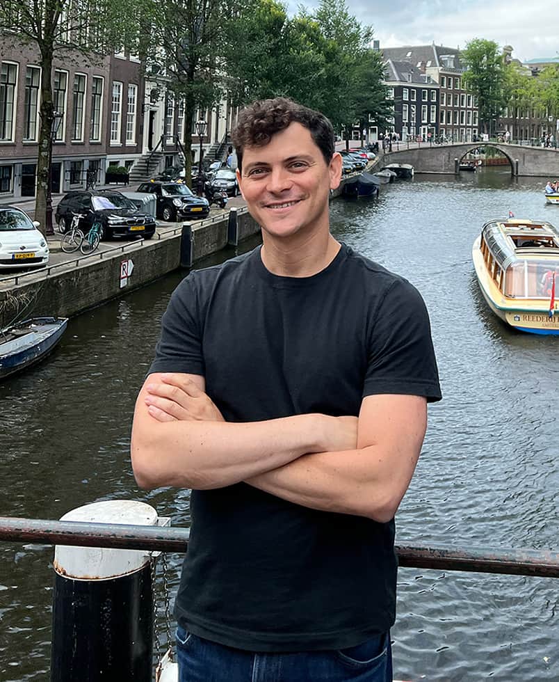 Nomadic Matt smiling with his arms crossed, wearing a black T-shirt, standing at a railing beside an Amsterdam canal with a tour boat and brick canal houses behind him.