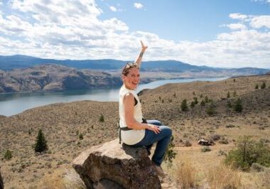 Woman on a British Columbia road trip sitting on a rock and smiling while pointing toward the scenic view from Battle Bluff Trail in Kamloops, with dry rolling hills, scattered pine trees, and Kamloops Lake shimmering under a bright blue sky.
