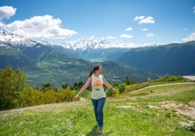 A happy female hiker walking down a grassy alpine ridge at the Cross Over Mestia viewpoint, showcasing expansive panoramic views of deep green valleys and towering, snow-capped Caucasus Mountain peaks in Svaneti, Georgia.