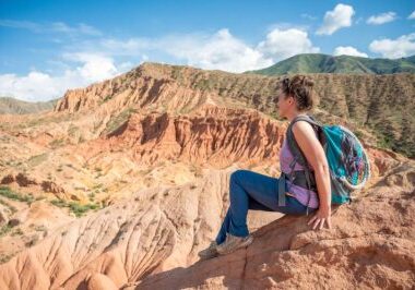 A woman sits on a rocky ledge in Fairytale Canyon, Kyrgyzstan, gazing out over the vibrant, eroded sandstone cliffs and ridges that stretch into the distance under a bright blue sky. She is wearing a backpack and casual hiking attire, capturing a moment of peaceful reflection amidst the canyon's stunning and colorful landscape.