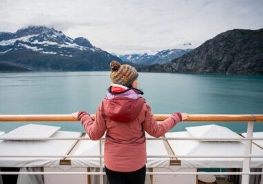 Traveler in a pink jacket and knit hat admiring the calm turquoise waters and dramatic mountain peaks of Glacier Bay National Park from the deck of a Holland America Alaska cruise ship.