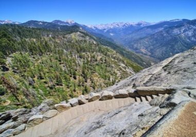 View looking down the steep stone staircase of the Moro Rock Trail in Sequoia National Park, California, with rugged granite cliffs and sweeping views of forested mountains and valleys in the distance under a clear blue sky.