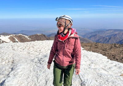 A female hiker walking on a snowy mountain ridge under a bright blue sky on the Mount Toubkal summit trail in Morocco. She is wearing green hiking pants, a pink winter jacket, a red scarf, a colorful knit hat, sunglasses, a climbing helmet with a headlamp, and crampons on her boots.