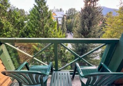 Private balcony at Pinnacle Hotel Whistler Village with two green chairs and a small table overlooking tall evergreens and distant mountains on a clear day.