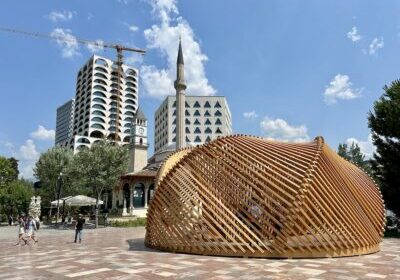 Modern wooden pavilion structure in Skanderbeg Square, Tirana, Albania, with the Et'hem Bey Mosque, clock tower, and modern high-rise buildings in the background on a sunny day.