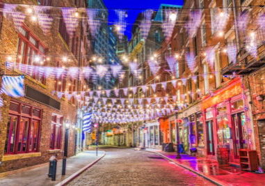 A vibrant nighttime view of Stone Street in Lower Manhattan, NYC, featuring cobblestone streets, historic brick buildings, and festive blue-and-white checkered bunting with string lights.
