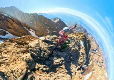 A wide-angle fisheye lens shot of a smiling female hiker sitting on the rocky edge of Mount Toubkal's summit. She is pointing up at the sky, wearing a white climbing helmet, red winter jacket, and green pants, with the Atlas Mountains curving along the horizon.